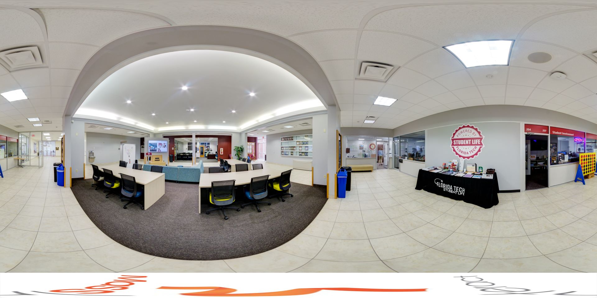 A panoramic view of a student life lounge at Florida Tech, featuring study tables with chairs, a lounge area, and various student service offices and informational displays.